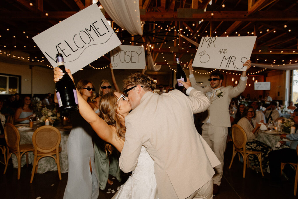 couple entering their reception with fun sunglasses and a bottle of champagne. Their wedding party was behind them, with big signs with their last name and their guests seated around them, clapping for them.